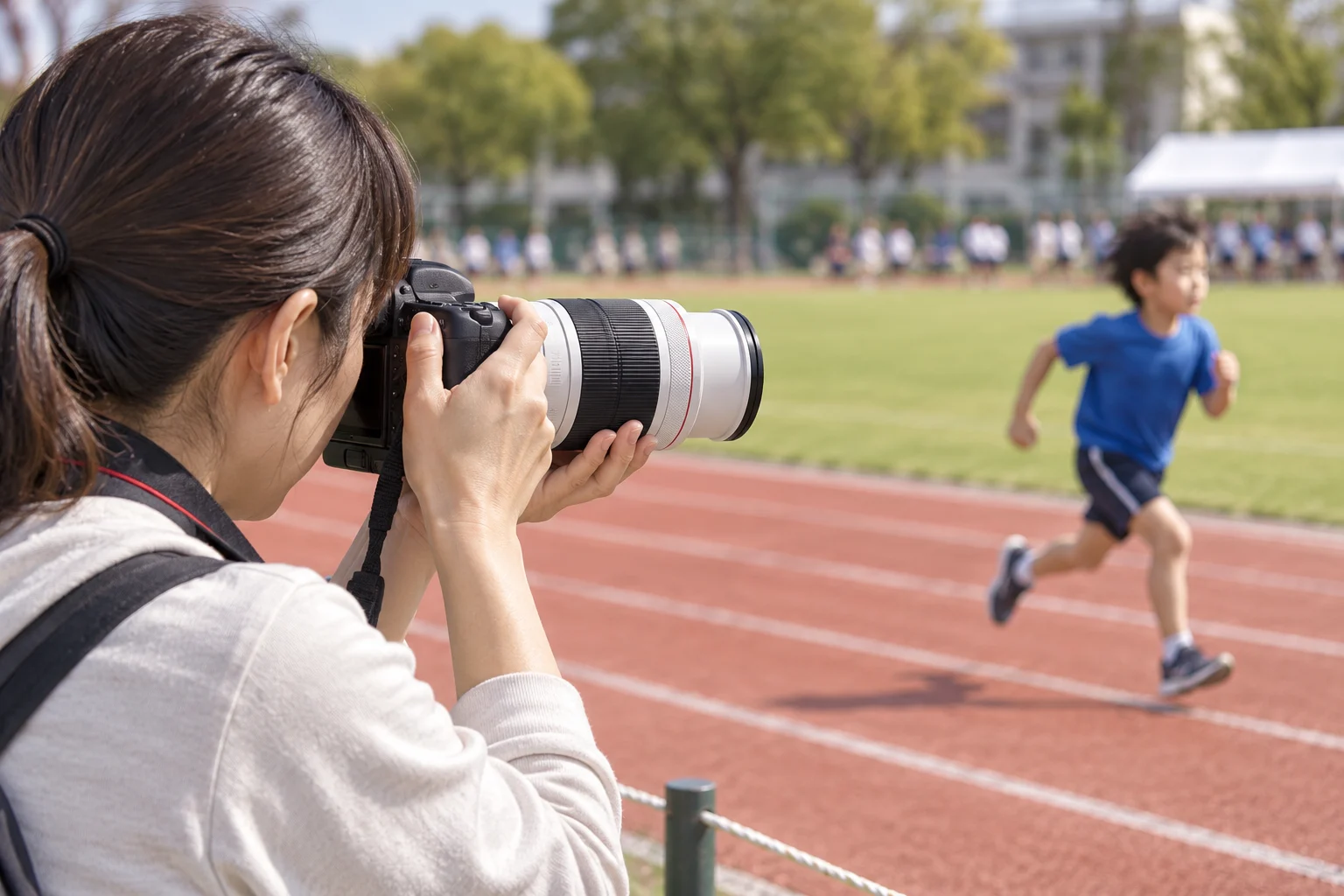 保護者席からRF70-200mm F4L付きカメラで青い服の走る子を撮る女性