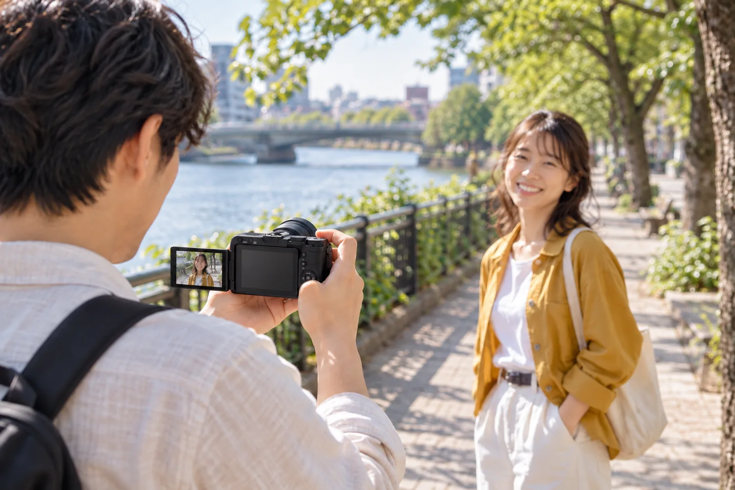 EOS R50 VとRF-S14–30mm PZを胸の高さに構える旅行者が、液晶を見ながら川沿いで笑う友人へレンズを向ける場面
