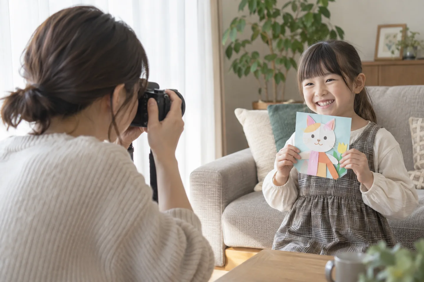 室内で工作を見せる子どもへ小さな単焦点レンズを向ける母親の撮影場面