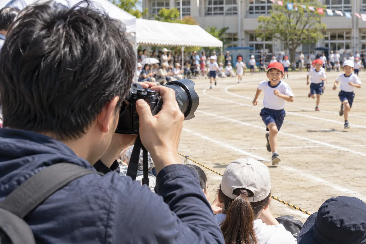 運動会の観客席から走る子どもへ望遠レンズを向ける父親の撮影場面